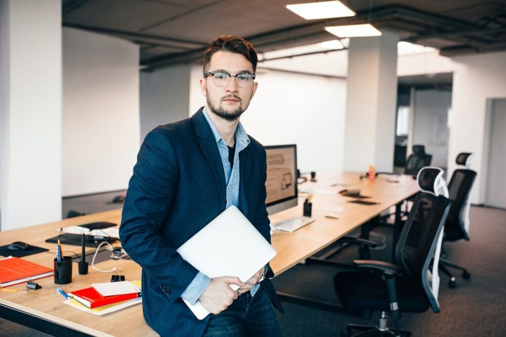 young-attractive-man-glassess-is-standing-near-workplace-office-he-wears-blue-shirt-dark-jacket-laptop-hand-he-is-looking-camera_197531-519